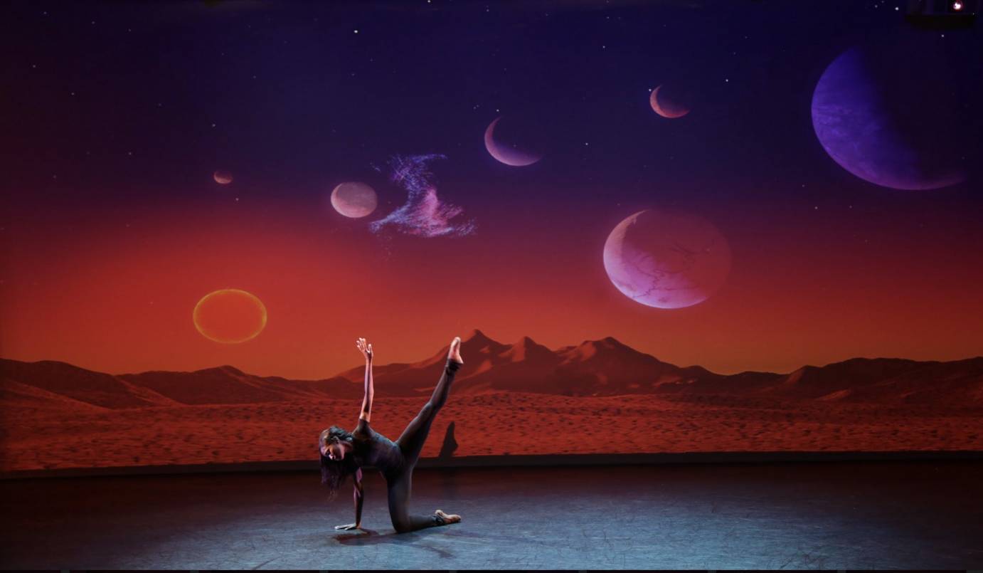 woman dressed in black balancing one palm and one knee in front of a backdrop of purple and magenta planets and moon