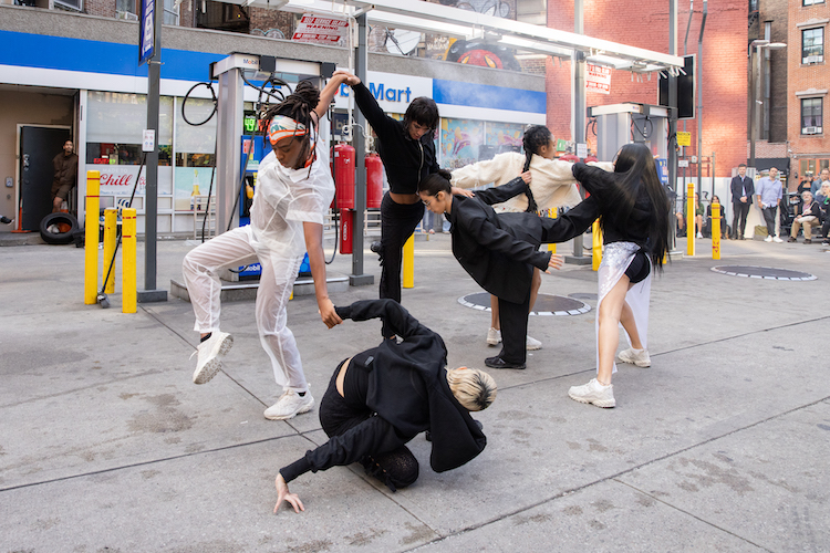 a group of dancers in various shades of white and black clothing hold hands in a circular dance in front of the Horatio Street gas station in NY