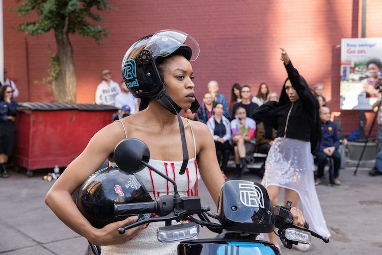 in the foreground a black woman on a motorcycle ponders holding her partners motorcycle helmet in the background her partner in a sheer skirt and black sweatshirt charges into space thrusting her hands in the air.