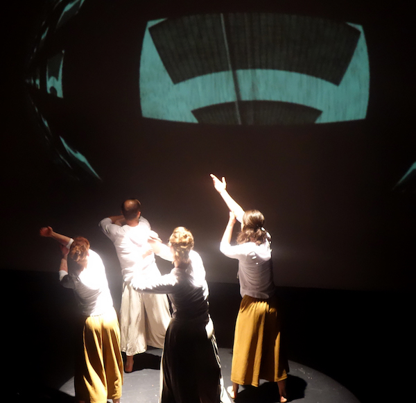 Four dancers in white shirts and yellow pants face the upstage wall which has a geometric projection
