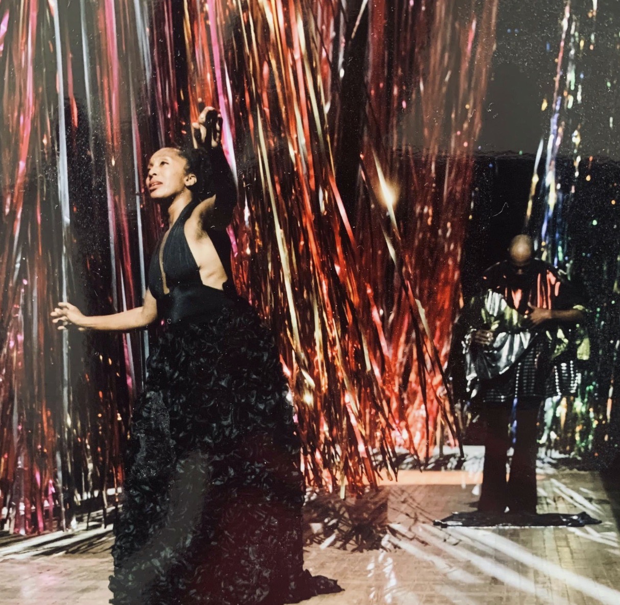 Francesca Harper in Foreground wearing a black halter gown with ruffled skirt.She is surrounded by large (ceiling to floor) strands of shiny tinsel-like material,there is a black man in the background of the shot with his head bowed.