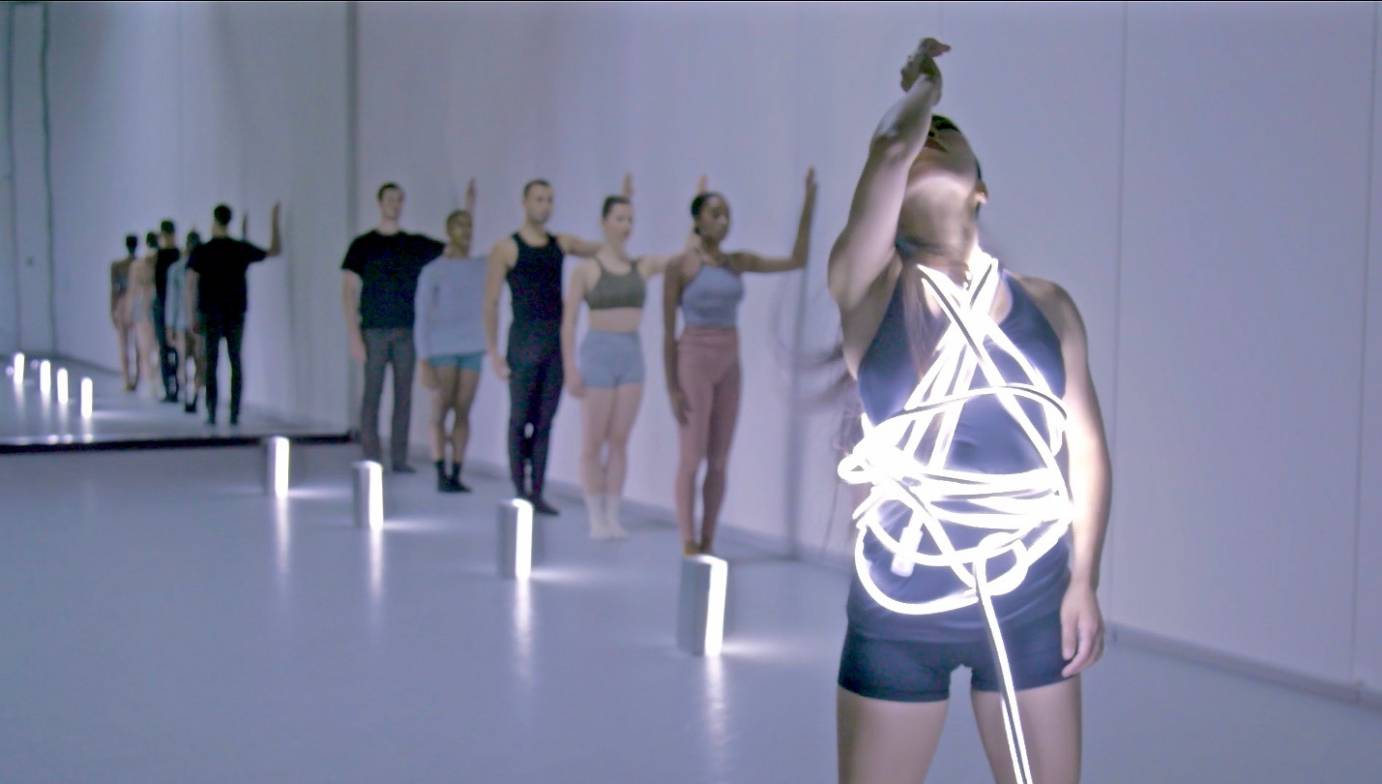 a company dancers in tight clothes of muted blues and tans stand in unison with their forearms against a white wall. In front of the groupis an Asian woman looking upwards her torso is encircled in ropes of light 