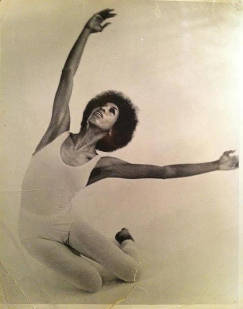 Denise Jefferson a black woman with an afro wearing an all white leotard ( sleevless, u-necked) and white tights, kneeling on the floor with arms in the air in the midst of a Graham modern dance exercise,the studio floor is white as is the background