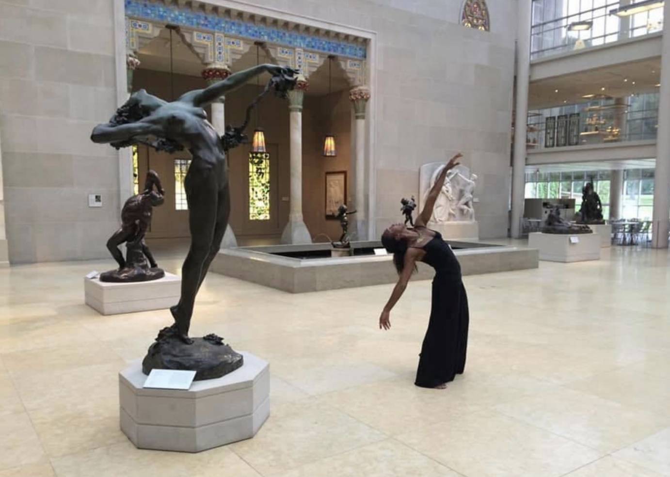 Francesca Harper a Black woman in a simple black gown with spagetti straps dances in the sculpture court of the Met. she is imitiating the pose of one of the statues leaning back with right arm extended to the sky. 