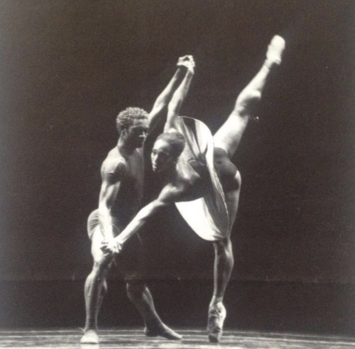 Desmond Richardson ( a muscled black man in a tshirt and shorts) supports Francesca Harper (a black woman in an ultra modern tutu) as she leans towards him on pointe, standing on one leg the other leg reaching high into the sky. the tutu is not netted but waves in a thin layer. the photo is black and white