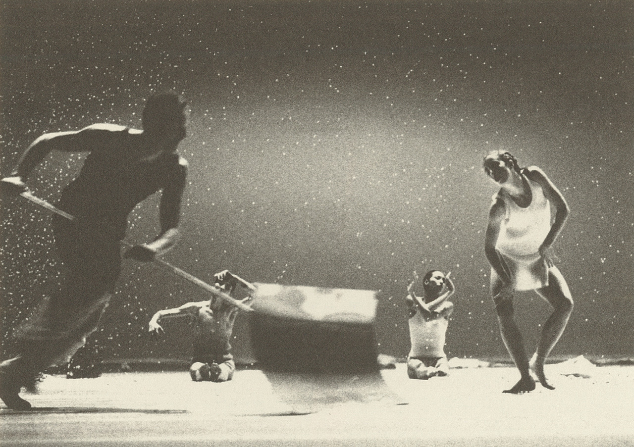 an old black and white picture, snow appears to be falling on stage ans one dancer a male to the left appears to be running with a snow shovel, two dancers a man and woman, he shirtless, she in a form fitting outfit,  sit on the floor gesturing their arms, another femal dancer leans to the left on a slightly bent leg with her right heel touching her left ankle, she wears a white sleevless top,and a sheer mini skirt,