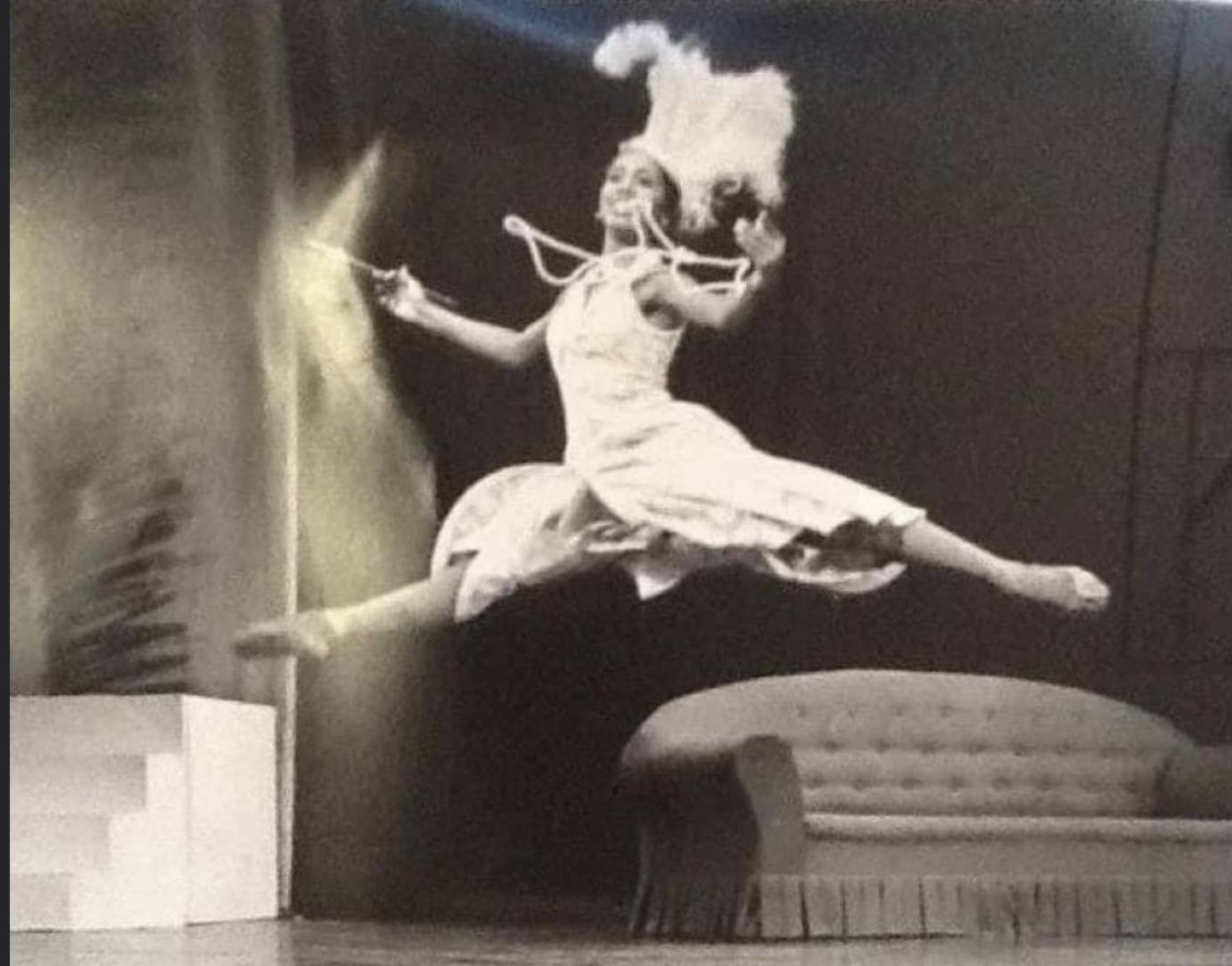 Francesca Harper a black woman, in grande jete ( big jump) wearing a feathered headress, a large strand of white pearls dances around her neck, and in one hand she holds a cigarette holdert 