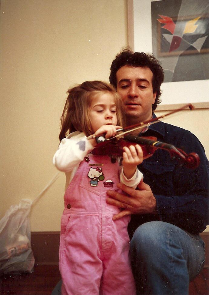 A father kneels beside his young daughter in pink overalls as she plays the violin