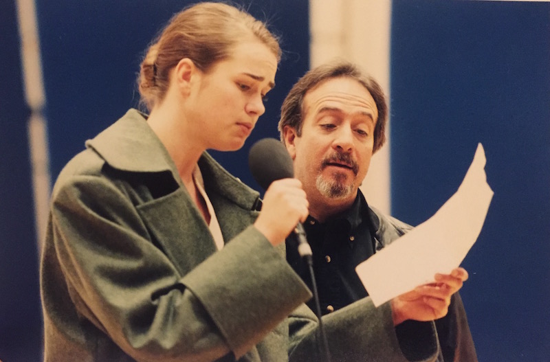 A bearded Michael Galasso stands next to a female performer who wears an overcoat and speaks into a microphone. They both are looking at a piece of paper.
