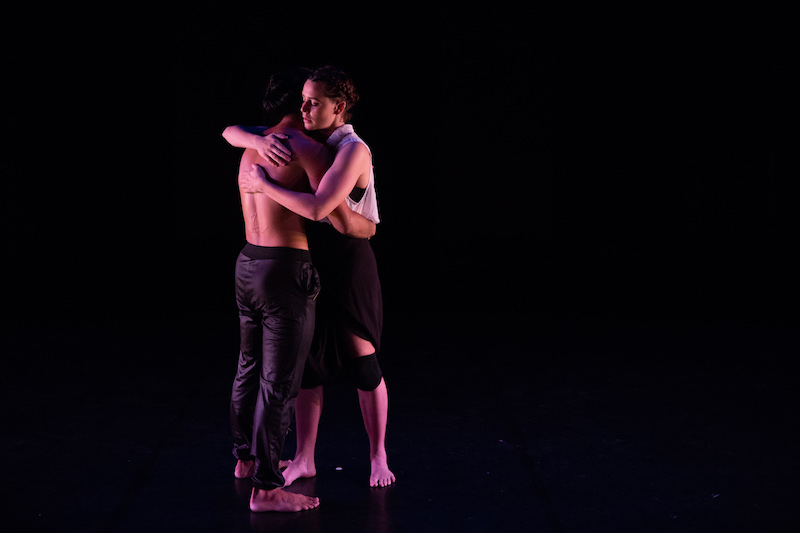 Two figures embrace. One is shirtless and other wears a white blouse and black bottoms.