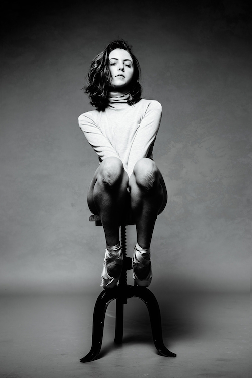 Gianna Reisen sits on a stool wearing a turtleneck and pointe shoes in a black and white photo.