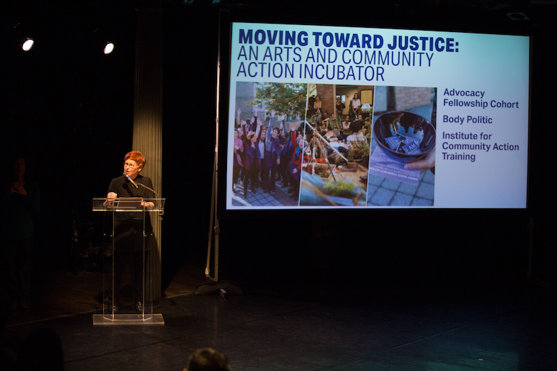 Gina Gibney stands at a podium describing the organization's new programming. A screen projections information about the new arts and community action incubator.