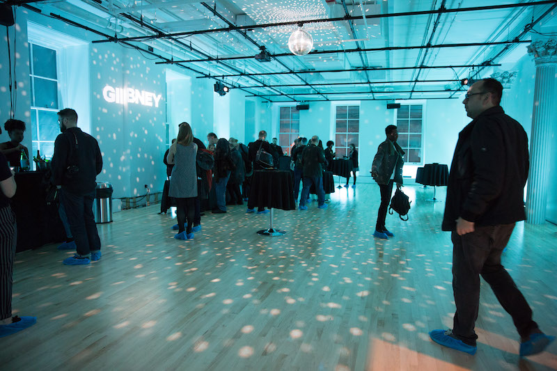 A crowd of people mingle in one of Gibney's new studios. A disco ball hangs from the ceiling casting an ice blue glow. The organization's new block-lettering logo 'GIBNEY' is projected on a wall