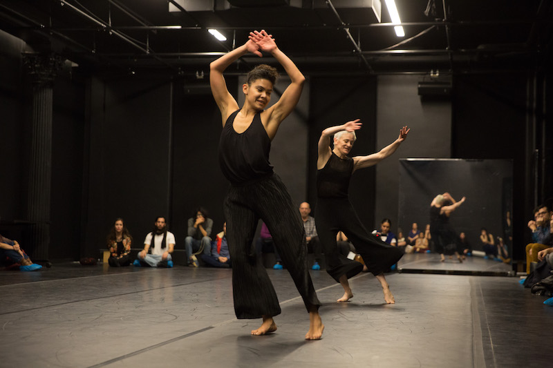 Two women in black tanks and pants dance with their arms above their heads. People sit around them and watch.