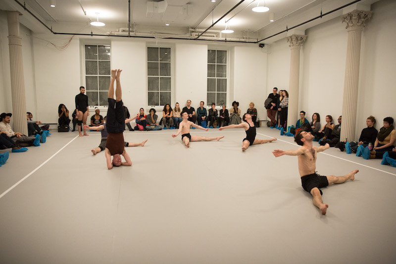 Dancers sit on the ground with their legs splayed. One stands on their head. Audience members line the studio's perimeter sitting and watch.