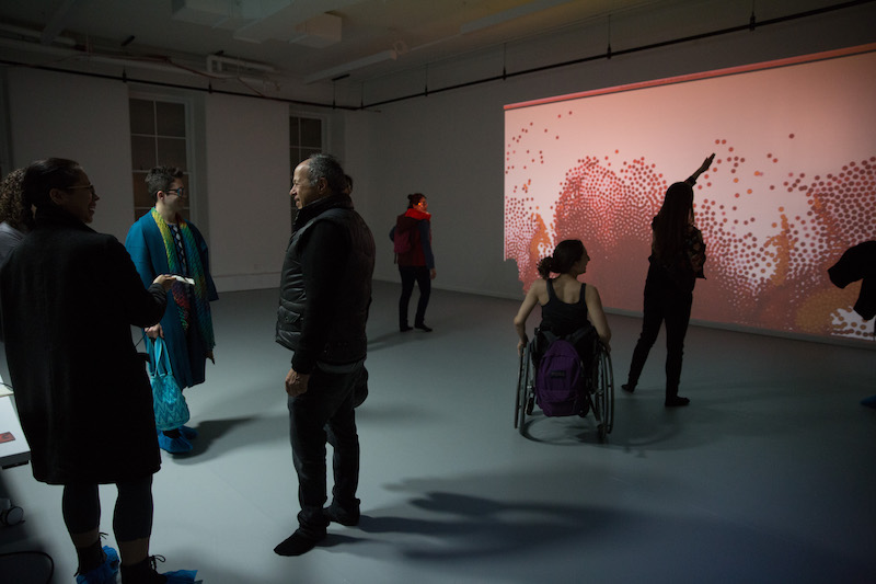 A group of people take in one of Gibney's new studios. A salmon colored screen with dots is projected on a wall.