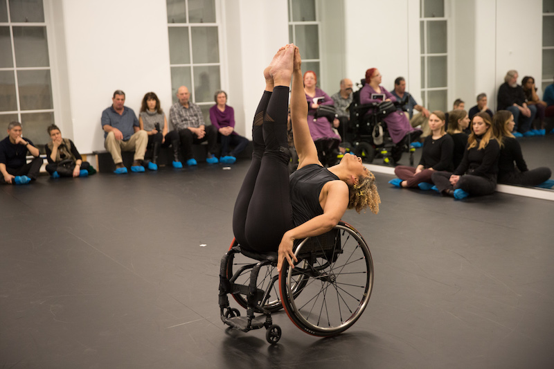 A woman raises her legs to sky as she extends back in a wheelchair. People sit on the floor and watch her in a Gibney studio.