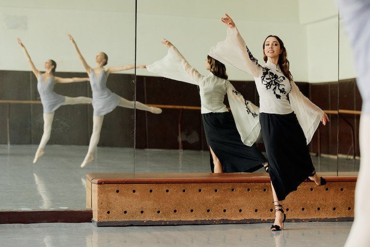 Kateryna Kukhar in an elegant white top embroiderd with black patterns, a long black skirt, and high heels, addressing her young students as we see them jump in the mirror 