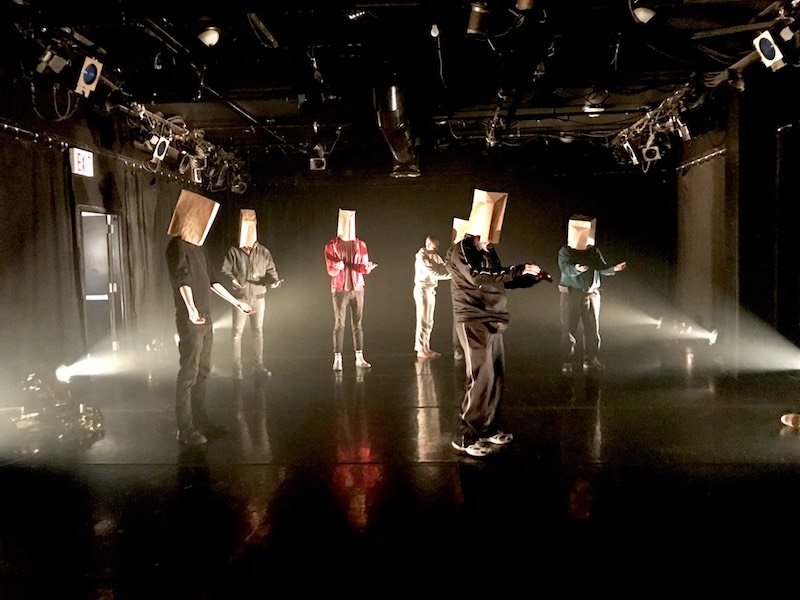 A group male audience members stand on stage with paper bags over their heads. Their arms are out in front of them.