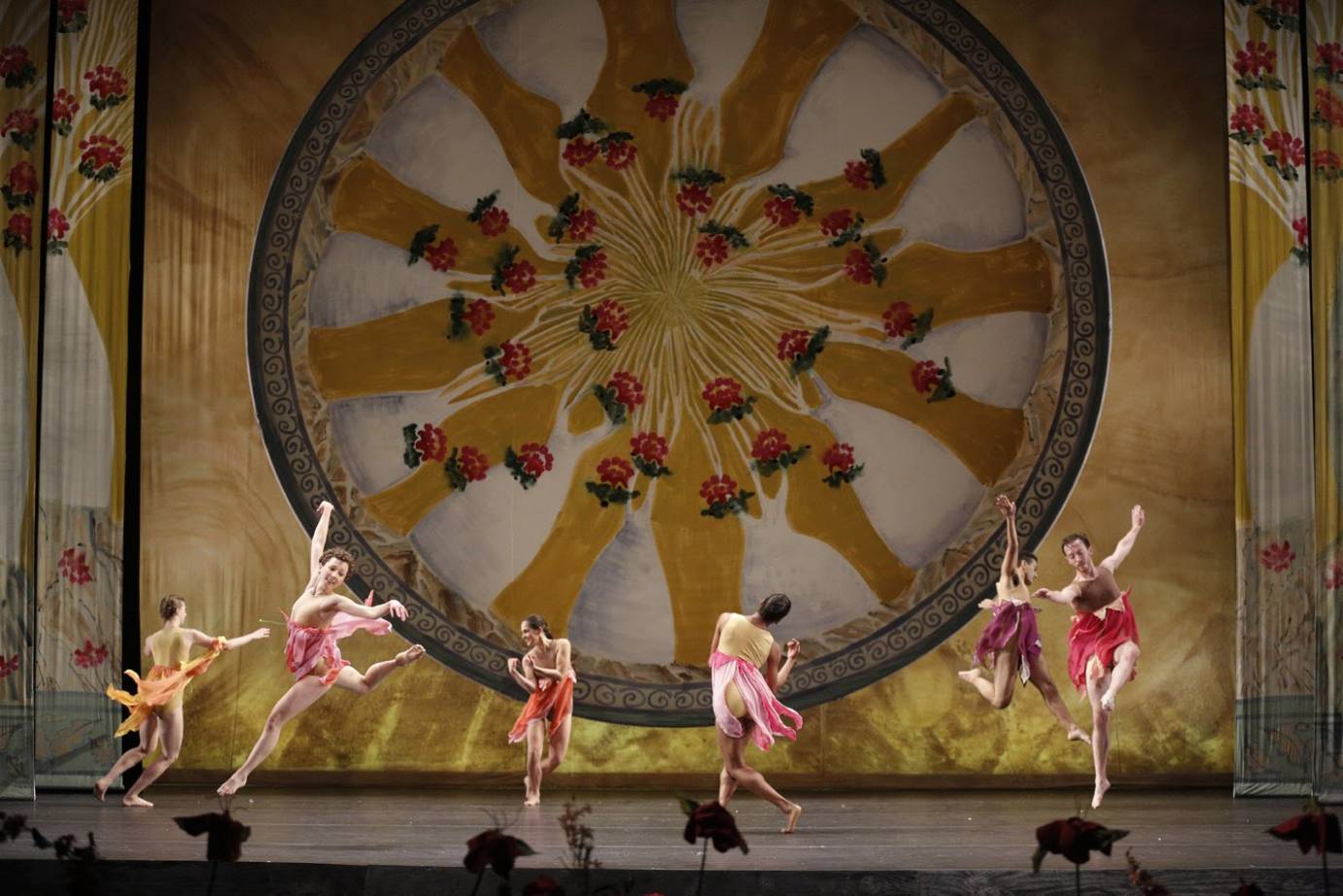 Six dancers in colorful costumes leap against an intricate gold background