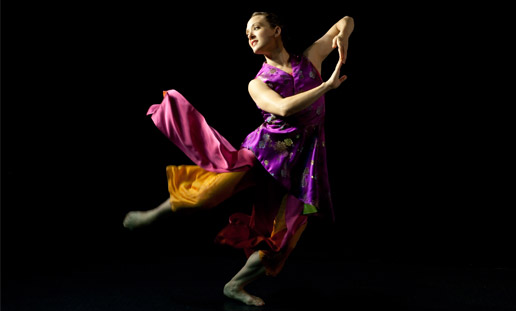 A woman in gem-colored tunic leaps into the air. Her arms bend at the elbow and her fingertips touch