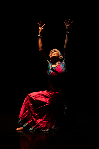 Bijayini Satpathy clothed in brigh red and blue gown kneels on the floor arms above her lifted to the sky, face filled with emotion to