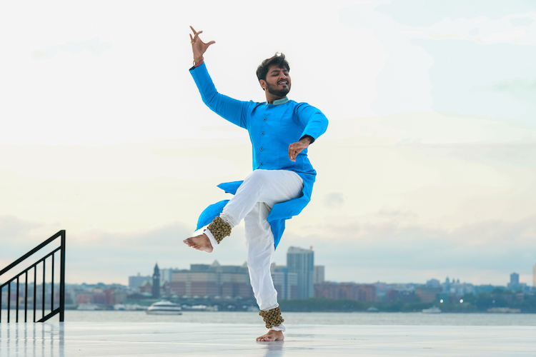 in a bright blue tunic and white pants with bells on his ankles a young bearded Indian man dances against the background of a New York sky