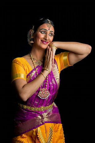 Portrait of an Indian dancer, Rasika Kumar, donned in bright yellow and purple with details of gold illuminating her outfit and face. Her  hands are pressed together palm of one pressing against the topsurface of another . She smiles, eyes wide. 