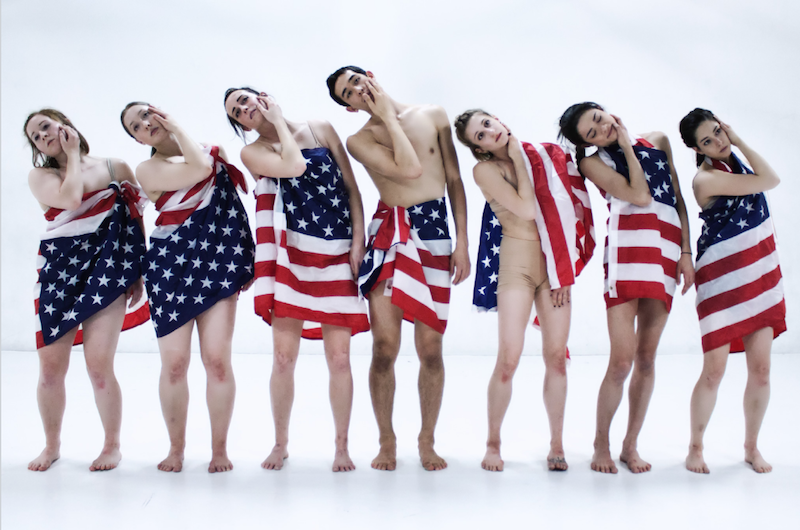 A group of dancers stand in a line with American Flags wrapped around their bodies. They each rest their right hand on their left cheek.