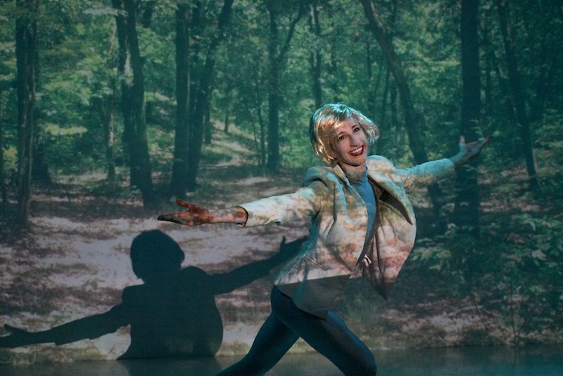 A woman in a blonde wig smiles and opens her arms in front of a forest projection.
