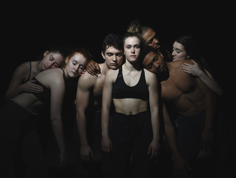 A group of dancers huddle in a group and lean on one another. They each close their eyes with the exception of one woman who stares directly at the camera.