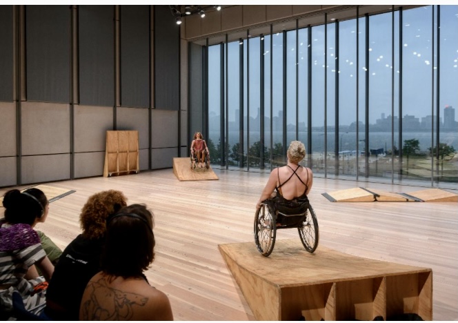 A wide shot: Laurel is in foreground, tipping backwards in their chair as they balance on a wooden ramp. Alice is across the studio, mirroring Laurel. A wall of tall windows is behind them and a few audience members, some wearing headphones, appear in the lower corner of the photo. Photo by Filip Wolak/ Whitney Museum of American Art. 