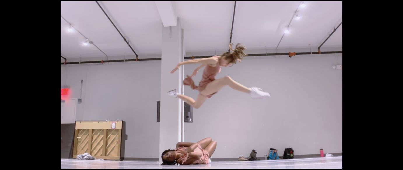 One female dancer stag leaps over a another dancer, who is curled on the floor