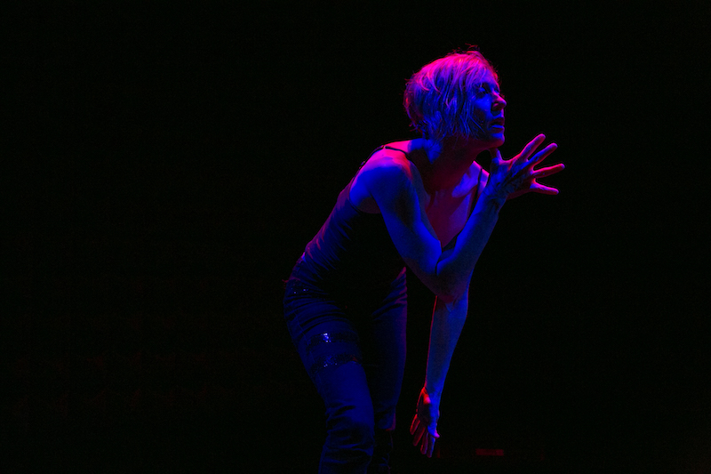Nicole Wolcott in rose-colored light gestures with her right hand, her fingers splayed, in front of her