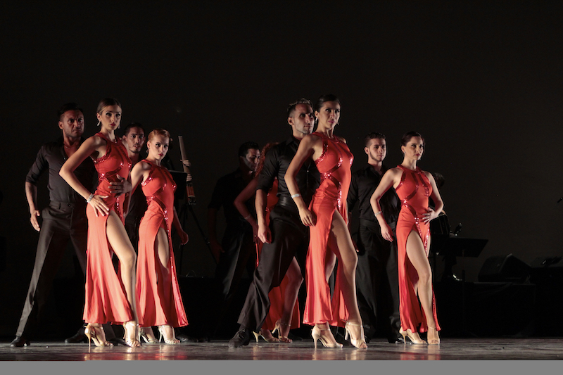 Tango dancers stand with their partners. The men wear all black and the women wear orange dresses with slits up the right thigh.