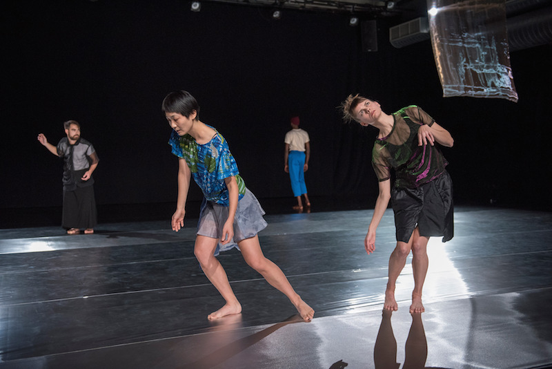 Two women in silk tops and skirts are in the background. Two others dancers stand in back. One wears a black filmy blouse and black skirt while the other faces the back curtain in blue pants and white tee.