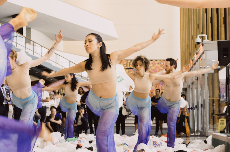 Dancers in purple tie dye pants and nude crop tops strike a pose with one leg bent behind them in a white lobby. Plastic bags are strewn about their feet. A crowd watches in the background.