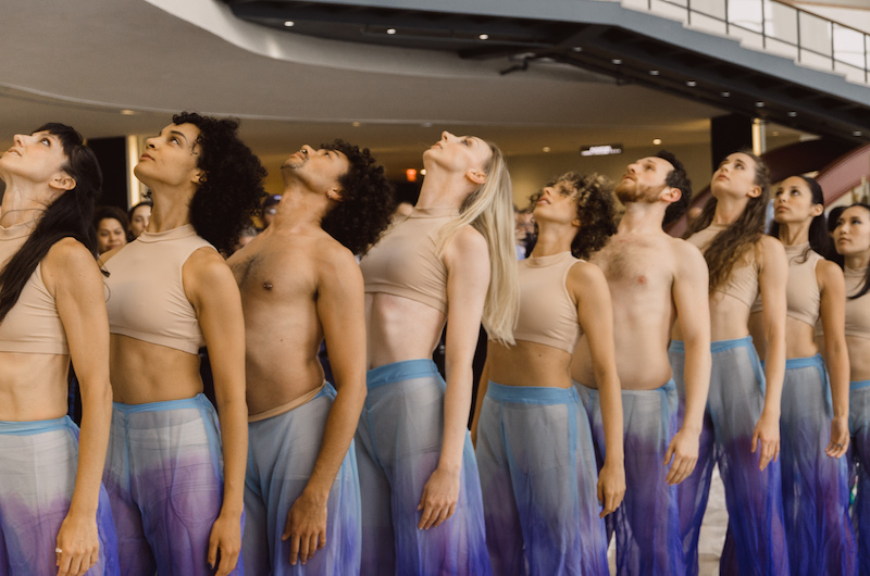 Dancers in purple tie dye pants and nude crop tops stand in a straight line and lift their head to the ceiling.
