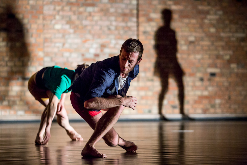 Two dancers cross their legs and crouch close to the ground. A shadow of a dancer standing is projected on the back wall