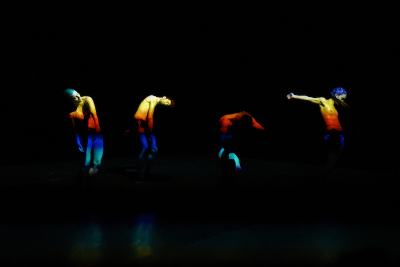 Four dancers stand in a line curving their torsos. A rainbow-colored light is projected on their bodies.