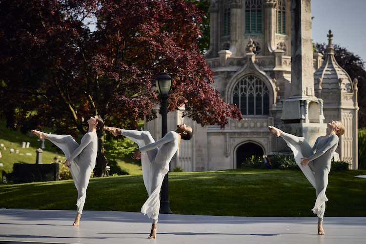 Three dancers in light blue unitards with sheer fabric skirts lean backward, suspended, poised on one leg with hands clasped around their extended leg.. the appearance is other worldly as the dancers perform against a backdrop of the Greenwood Cemetery and its chapel