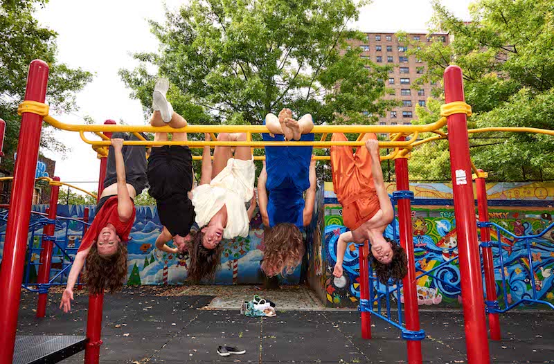 The five members of the Lovelies hang upsidedown from yellow monkey bars in an outdoor playground