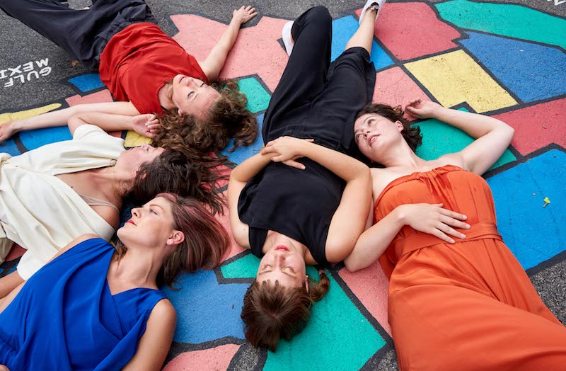 Five women in in colorful contemporary clothes lay atop a pavement mural of the 50 states.