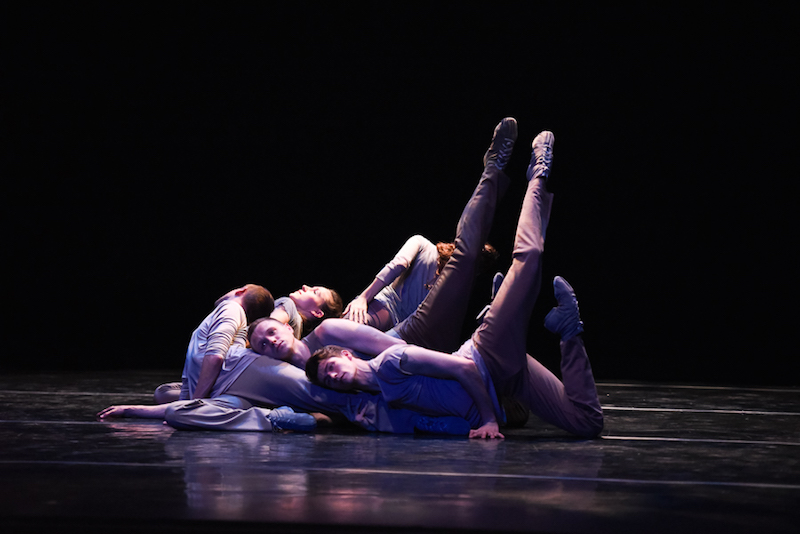 Dancers lay on one another in cascading heap on stage. Two dancers lift their legs in attitude positions above their heads.