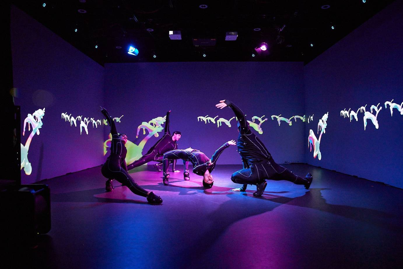 Three dancers in black, low to the ground with one leg bent and the other extended, surround a dancer in the center performing a backbend. The dancers' images, transformed into little meme-like figures, are projected on the purple wall. 