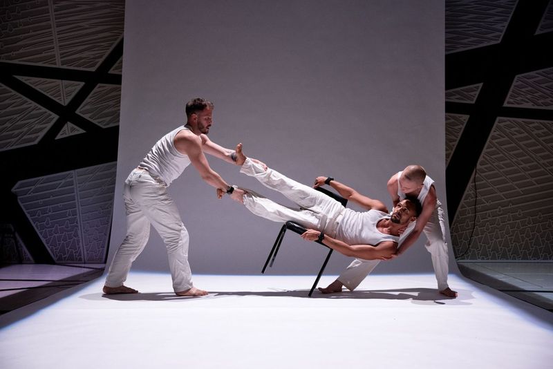 Three dancers in white: one lying prone across a chair pulled by the feet by a dancer and and whose shoulders and head are nestled into the arms of a dancer.