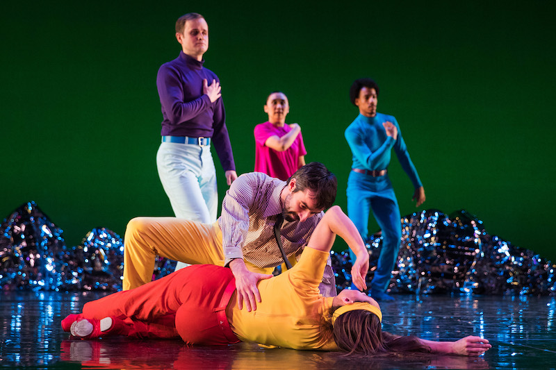 Three dancers in neon hues cross their hands over their hearts. While a couple in the foreground crouch to the floor.