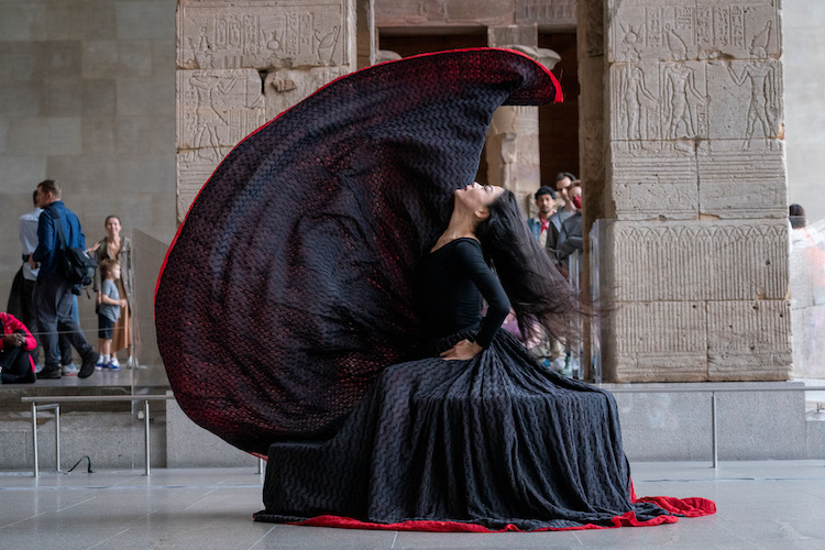 a woman seated on a bench in profile flings her prodigious skirt in the air ..the colors of her voluminous skirt are striking andbold black with a blood red underskirt 