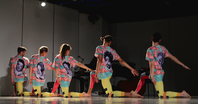 A group of dancers in color leggings and tees kneel in a straight line. their right arms extend on a low diagonal.