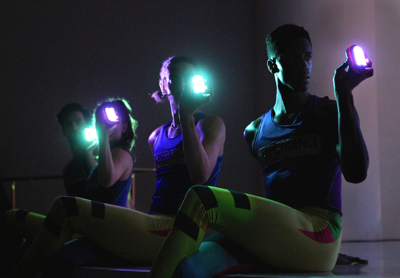Three dancers in blue tank tops and yellow neon leggings shine lights in front of their faces